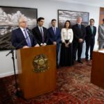 Fotografia de uma cerimônia oficial em ambiente interno. Uma pessoa fala ao microfone atrás de um púlpito de madeira com o brasão nacional, enquanto sete pessoas permanecem alinhadas ao fundo. À esquerda, há uma bandeira do Brasil. À direita, uma mesa de madeira exibe documentos para assinatura. O ambiente possui tapete estampado em tons de vermelho, paredes claras e grandes fotografias em preto e branco penduradas ao fundo. Luz natural entra pelas janelas localizadas no lado direito da sala.