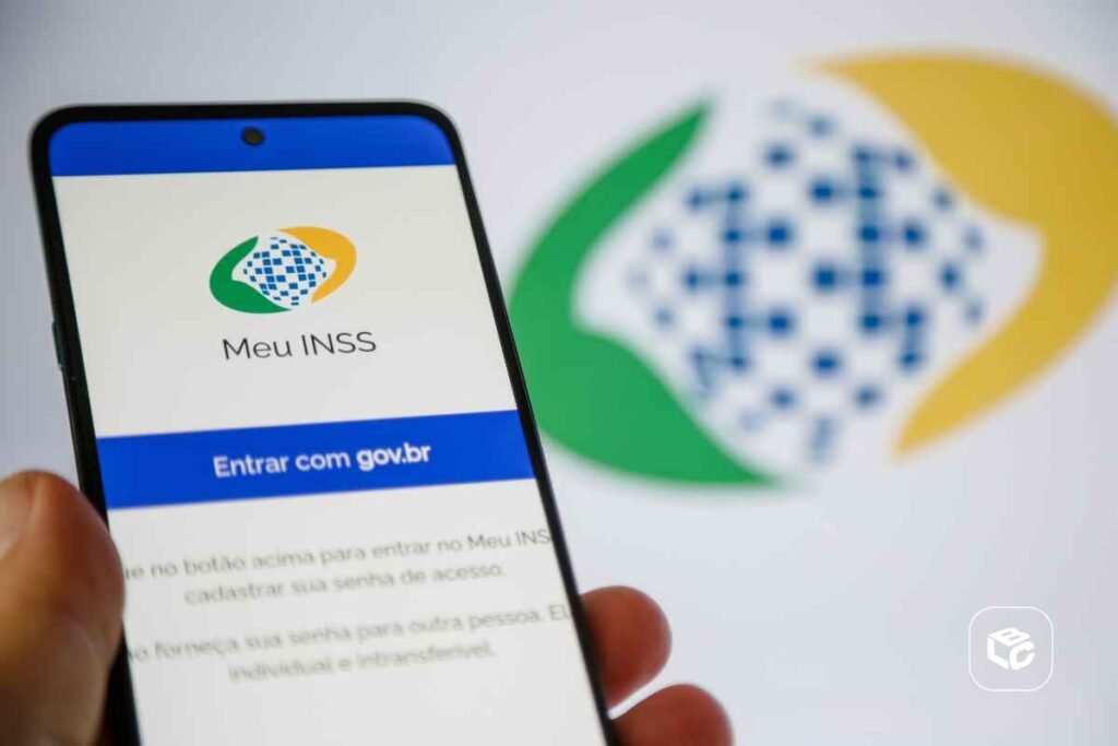 Smartphone exibindo a tela inicial do aplicativo Meu INSS, com o logotipo da plataforma e o botão azul “Entrar com gov.br”. O aparelho é segurado por uma mão em primeiro plano. Ao fundo, desfocado, aparece o logotipo do INSS em verde, amarelo e azul sobre um fundo claro. A imagem destaca o acesso digital aos serviços da Previdência Social por meio de dispositivo móvel.