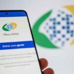 Smartphone exibindo a tela inicial do aplicativo Meu INSS, com o logotipo da plataforma e o botão azul “Entrar com gov.br”. O aparelho é segurado por uma mão em primeiro plano. Ao fundo, desfocado, aparece o logotipo do INSS em verde, amarelo e azul sobre um fundo claro. A imagem destaca o acesso digital aos serviços da Previdência Social por meio de dispositivo móvel.