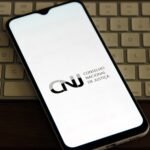 Smartphone com a tela exibindo o logotipo e o nome “CNJ - Conselho Nacional de Justiça”, apoiado diagonalmente sobre um teclado de computador claro. O aparelho está sobre uma superfície de madeira escura, com foco na tela iluminada e fundo levemente desfocado.
