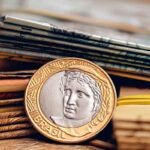"Moeda brasileira de 1 real posicionada em primeiro plano sobre uma pilha de cédulas de real. A imagem destaca detalhes da moeda e das notas ao fundo, com foco seletivo e iluminação quente que remete a finanças, economia e dinheiro."