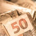 Close-up de cédulas de real brasileiro empilhadas, com destaque para uma nota de R$ 50 em primeiro plano. A imagem apresenta foco seletivo, deixando partes das demais notas desfocadas ao fundo. Tons quentes predominam na composição, remetendo a finanças, recursos financeiros, pagamentos e valores monetários. Logotipo da empresa visível no canto inferior esquerdo.