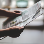 Mãos segurando e lendo um jornal aberto em ambiente interno. O enquadramento destaca o jornal e as mãos em primeiro plano, enquanto o restante do ambiente aparece desfocado ao fundo. Parte das páginas exibe textos e imagens impressas, mas o conteúdo não está legível devido ao desfoque. A iluminação é suave e natural, criando contraste entre o jornal e o fundo em tons neutros. Um logotipo azul está visível no canto superior direito da imagem.