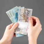 Mãos segurando e contando cédulas de real brasileiro, incluindo uma nota de R$ 200 e outras notas azuis, sobre fundo cinza liso.